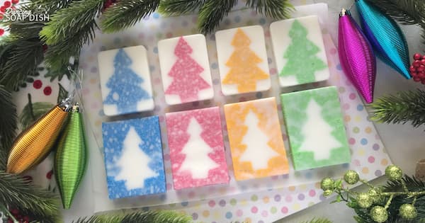 Eight rectangular soaps arranged in two rows show colorful Christmas tree designs made from exfoliating cellulose sponge. The top row has white bars with blue, pink, orange, and green sponge trees, while the bottom row reverses the colors with white trees on bright sponge backgrounds. They sit on a white tray surrounded by pine branches and shiny ornaments in green, gold, purple, and blue.