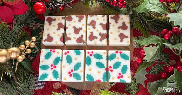 Eight rectangular soaps arranged in two rows sit on a festive surface decorated with holly leaves, red berries, pine branches, and gold ornaments. The top row features white bars patterned with brown reindeer heads with bright red noses, while the bottom row shows white bars decorated with holly leaves and berries.