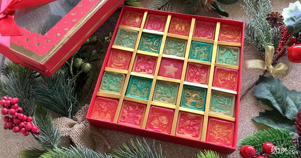 A festive gift box holds twenty‑five square red and green soaps arranged in a 5×5 grid, each embossed with different Christmas designs and numbered 1-24. A red square soap with a gold star sits in the centre of the box. Golden dividers separate the pieces, and pine branches, holly berries, and ribbons surround the box, creating a cheerful Christmas display.