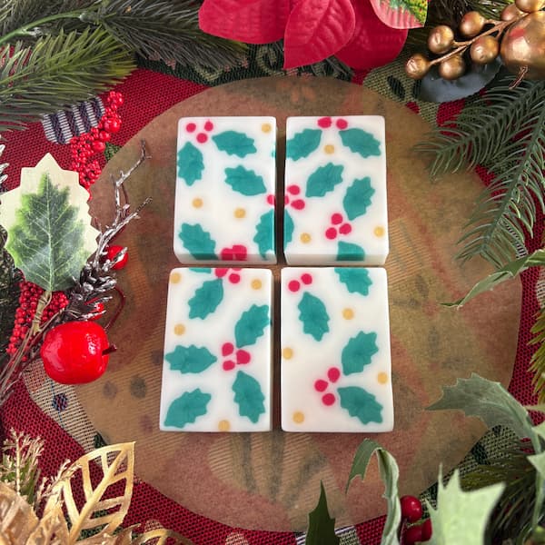 Four rectangular white handmade soaps decorated with green holly leaves, red berries, and small gold dots are arranged in a grid. The soaps are surrounded by festive Christmas greenery, pine needles, red berries, gold leaves, and a bright red poinsettia flower, creating a traditional holiday-themed display.