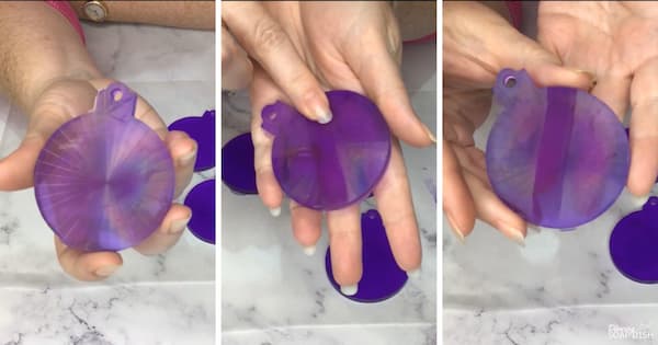 A three‑panel collage showcases translucent purple ornament‑shaped soaps. The soaps display shifting swirls and bands of purple, pink, blue, and lavender that catch the light, creating an iridescent, holographic appearance. Each panel shows a hand holding a circular soap ornament, with the second panel also showing a finger running across the surface of the soap, wiping away the holographic pattern as it does so.