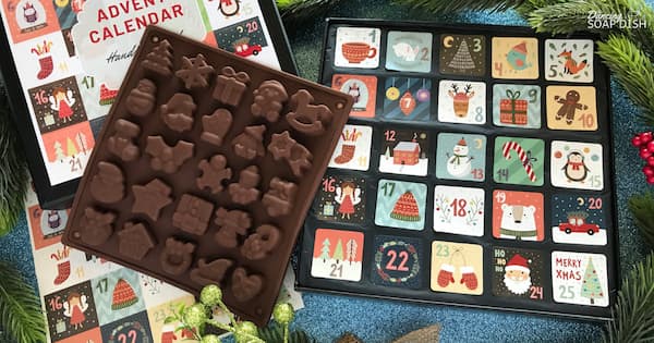 A Christmas‑themed advent calendar set is displayed among evergreen branches and red berries. On top lies a silicone mould of small festive shapes such as stars, gifts, snowmen, gingerbread men, bells, and trees. Beside it sits a colorful advent calendar with numbered compartments from 1 to 25, each decorated with holiday illustrations like Santa, mittens, ornaments, and reindeer.