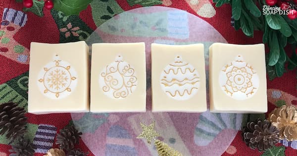 Four rectangular soaps with Christmas ornament embeds embossed with various gold designs, a snowflake, a swirl, wavy lines, and a floral motif, are arranged on a festive red‑and‑green background with pinecones, greenery, and a small gold star.