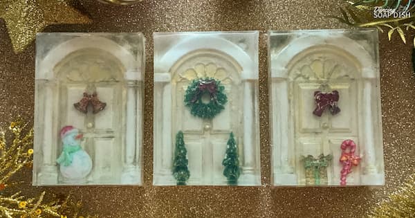 Three rectangular soaps are arranged on a glittery gold surface, each designed like a festive front door. One features a pair of red bells with a snowman, another shows a green wreath with tiny Christmas trees, and the third has a red bow with presents and a candy cane. Decorative gold foliage surround the display.